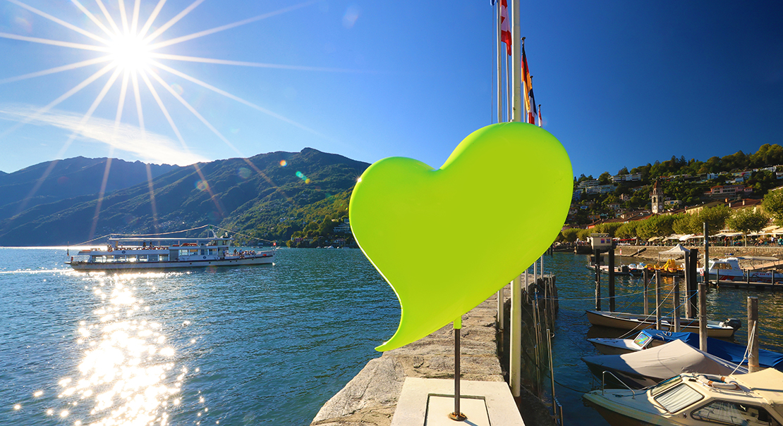 Green heart sculpture on the Ascona waterfront beside a sunlit lake and passing boat