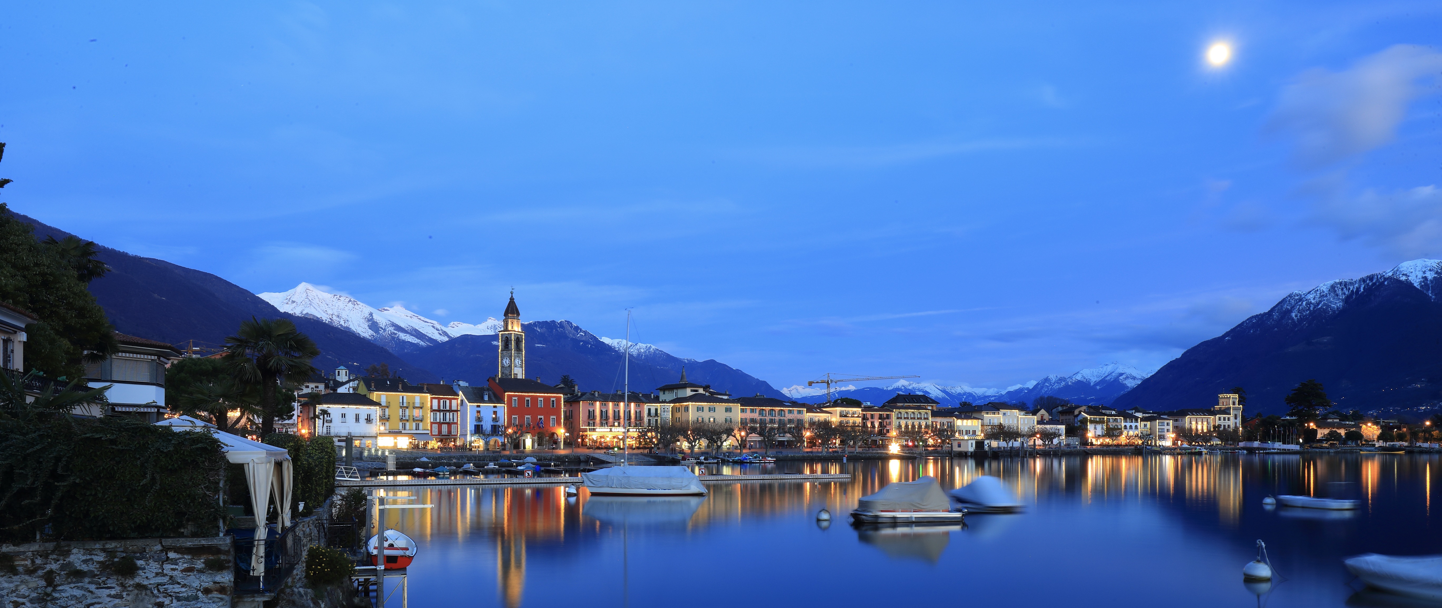 Evening harbor of Ascona with boats, church tower, and distant snowy mountains