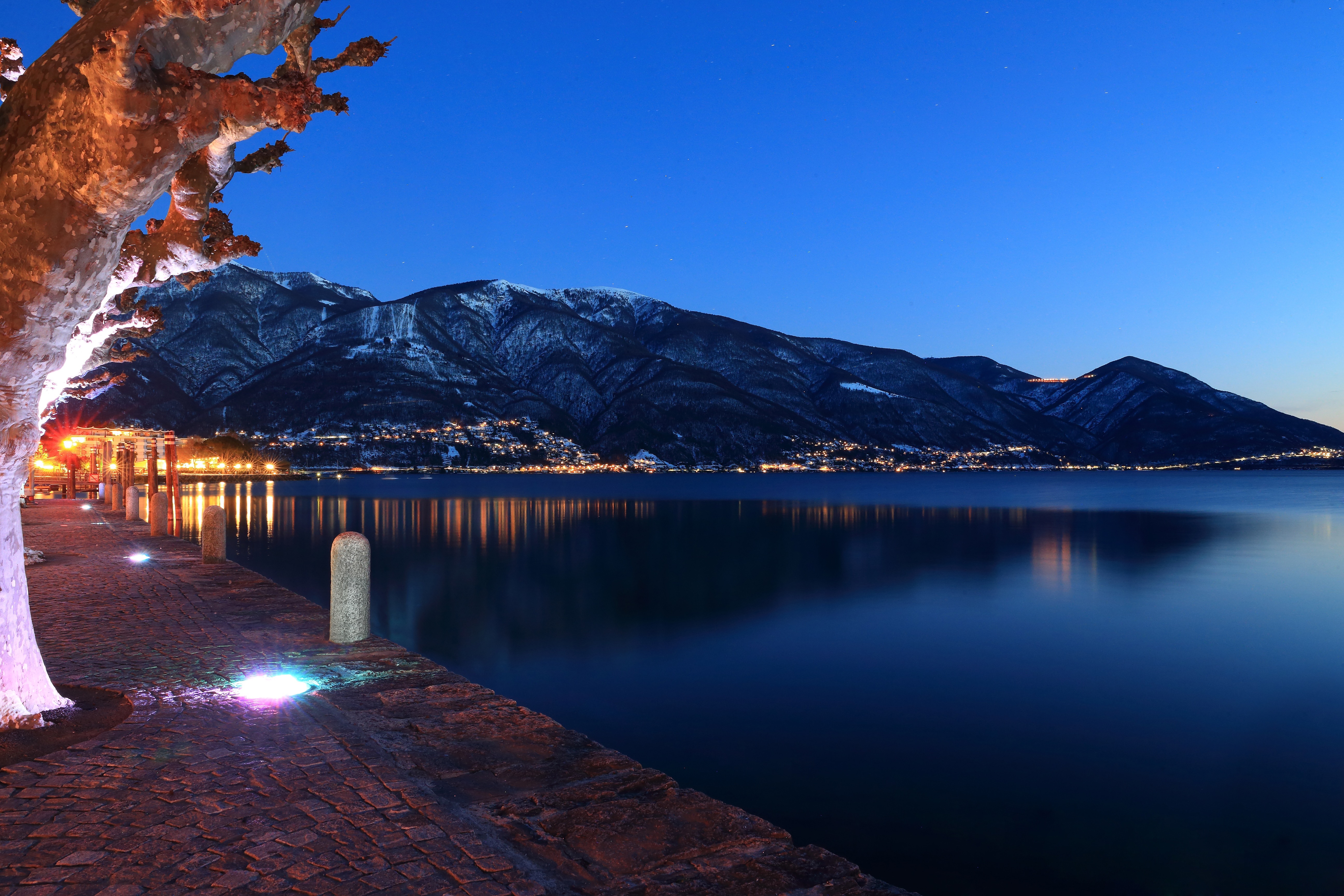 Blue-hour lake promenade with lit walkway and snow-dusted peaks
